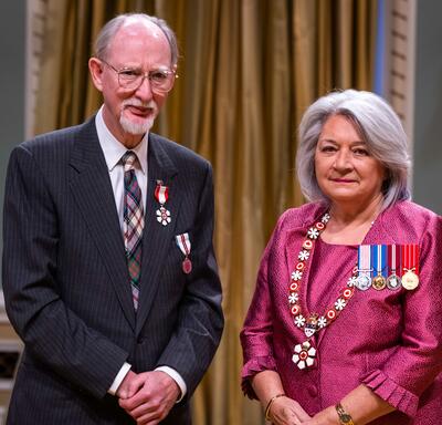 Governor General Simon is standing next to a man who has a medal pinned to his grey coat.