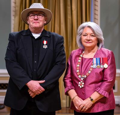 Governor General Simon is standing next to a man who has a medal pinned to his black blazer. He is wearing a hat.