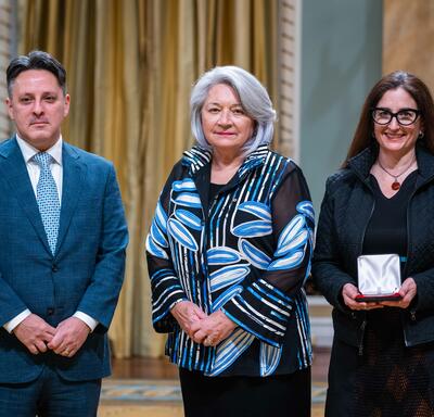Governor General Simon is standing in between a man and a woman. The woman is holding a small box with a medallion inside of it.