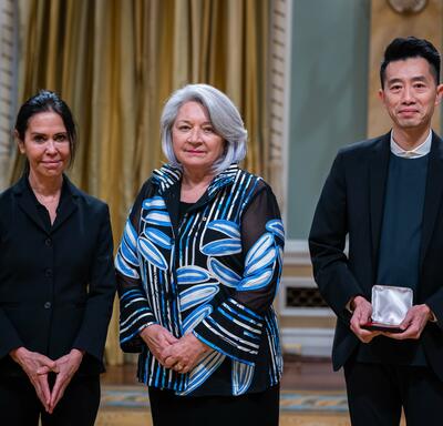 Governor General Simon is standing in between a man and a woman. The man is holding a small box with a medallion inside of it.