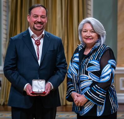 Governor General Simon is standing next to man. The man is holding a small box with a medallion inside of it.