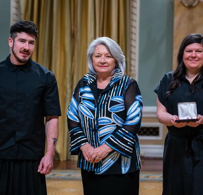 Governor General Simon is standing in between a man and a woman. The woman is holding a small box with a medallion inside of it.