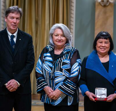 Governor General Simon is standing in between a man and a woman. The woman is holding a small box with a medallion inside of it.