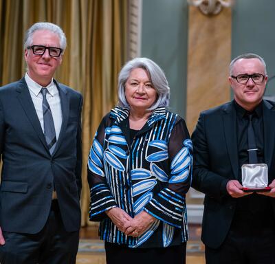 Governor General Simon is standing in between two men. The man on the right is holding a small box with a medallion inside of it.