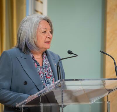 Governor General Simon is speaking into a microphone. She is wearing a blue-grey blazer with a colourful blouse.