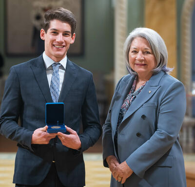 Governor General Simon is standing next to a young man. He is holding a blue box with a medallion inside of it.