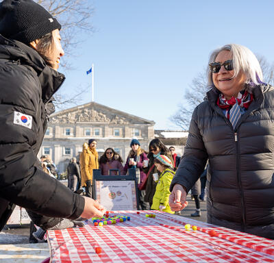 Governor General Simon speaks to a person standing behind a table. There is a colourful game on the table. They are outdoors and wearing winter clothing. 