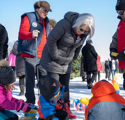 Governor General Simon bends over to speak to several children playing in the snow. Mr. Fraser and several others stand around and behind her.