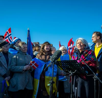 Governor General Simon speaking into a microphone. A crowd is gathered around her. Many people are holding flags from different countries.