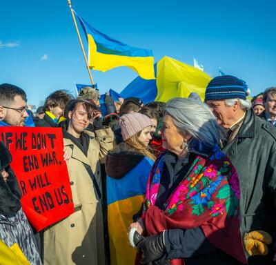 Governor General Simon and Mr. Fraser walk through a crowd of people. She looks at a man holding a red sign that reads, “If we don’t end war, war will end us.” Many other people are holding Ukrainian flags.