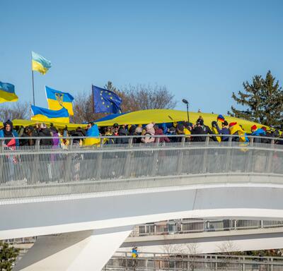 A group of people stand on a bridge. They are holding a large, yellow-and-blue Ukrainian flag above them. Others are waving smaller Ukrainian flags. A line of trees and blue sky are behind them.