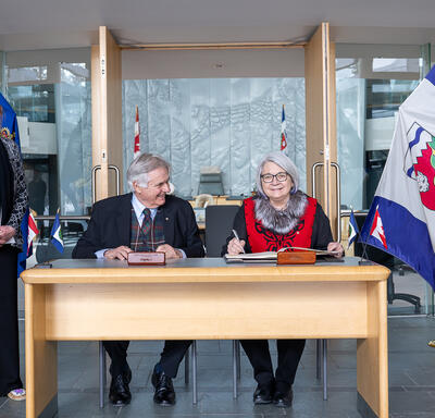Governor General Simon and Mr. Fraser are sitting at a wood table. The Honourable Caroline Cochrane is standing to their right, and is smiling at the camera. 
