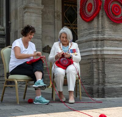Governor General Mary Simon crocheting with another person. They are seated outside of Rideau Hall.
