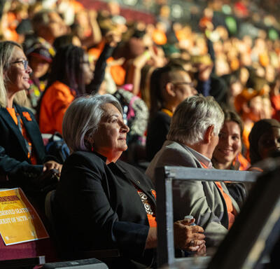 The Governor General sitting in a large stadium with hundreds of people. Many are wearing orange.
