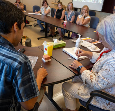 Un groupe de personnes est assis autour d'une table de conférence. Une femme aux cheveux gris tient une seringue tandis que les autres la regardent attentivement.