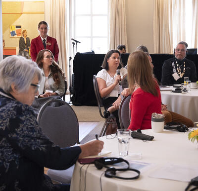 Un groupe de personnes assises autour de tables rondes. Une personne tient un micro.