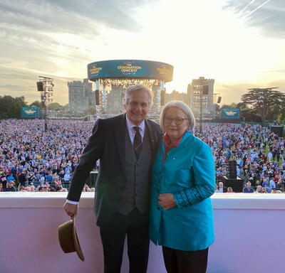 Governor General Simon and Mr. Whit Fraser standing on a balcony posing for a photo. The balcony overlooks a large crowd of people. Behind them in the distance is a large stage. 