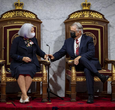 Governor General Mary Simon and Mr. Whit Fraser sitting in throne seats, holding hands.