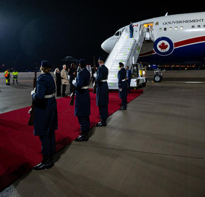 The Government of Canada plane is on the tarmac at the airport. The door to the plane is open and the stairs are open waiting for the passengers to descend. There is a red carpet at the base of the plane.