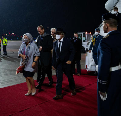 Governor General Mary Simon and Mr. Whit Grant Fraser are greeted by German and Canadian officials. They are on a red carpet at the foot of a plane. There are people in military uniform along the red carpet. It is nighttime. 