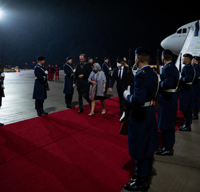 Governor General Mary Simon and Mr. Whit Grant Fraser are greeted by German and Canadian officials. They are on a red carpet at the foot of a plane. There are people in military uniform along the red carpet. It is nighttime. 