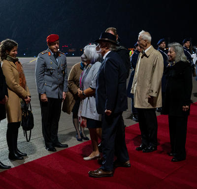 Governor General Mary Simon and Mr. Whit Grant Fraser are greeted by German and Canadian officials. They are on a red carpet at the foot of a plane. There are people in military uniform along the red carpet. It is nighttime. 