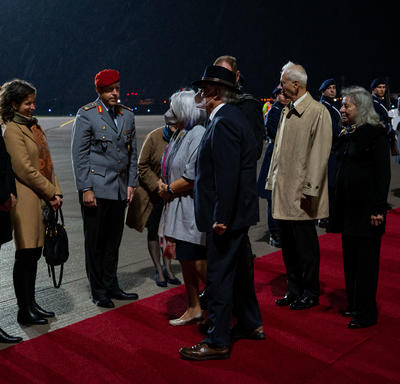 La gouverneure générale Mary May Simon et Monsieur Whit Grant Fraser sont accueillis par des représentants allemands et canadiens. Ils sont sur un tapis rouge au pied d’un avion. Des personnes en uniforme militaire sont debout le long du tapis rouge.