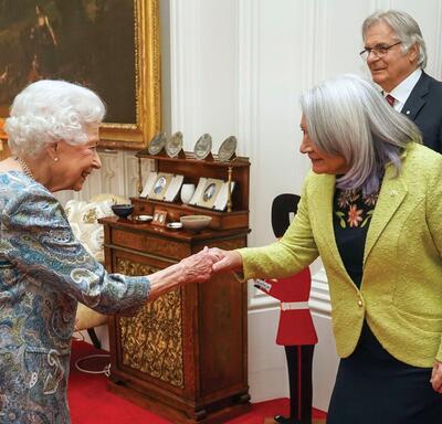 Governor General Mary Simon shaking hands with Queen Elizabeth II. Behind them, Mr. Whit Fraser stands and smiles.