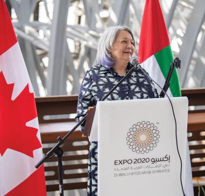 Governor General Mary Simon standing at a podium and delivering remarks. The national flag of the United Arab Emirates is on display behind her. It is red, green, white, and black.