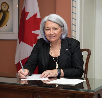 Governor General Mary Simon sitting and signing a paper document at a desk. A Canadian flag is on display behind her.