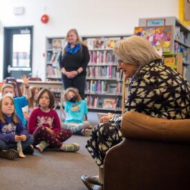 La gouverneure générale Simon fait la lecture à un groupe d'enfants assis devant elle dans une bibliothèque. Elle est assise dans un fauteuil marron. Une enseignante se tient debout derrière le groupe.