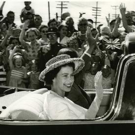 Photo en noir et blanc d’une jeune reine Elizabeth II. Elle salue la foule depuis le siège arrière d’une voiture décapotable. Elle porte un chapeau, un collier de perles et des gants.