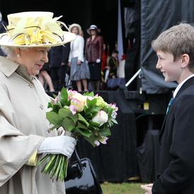 Queen Elizabeth II is wearing a wide-brimmed hat with yellow flowers and a light-tan-coloured trench coat. She holds a bouquet of flowers. A young boy stands in front of her. The Duke of Edinburgh trails behind her. 