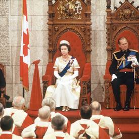 Queen Elizabeth II and the Duke of Edinburgh are seated in a pair of thrones. The Queen is wearing a cream-coloured dress, a crown and a navy blue sash. The Duke is wearing a full military uniform. A Canada flag is to the Queen’s immediate right. 