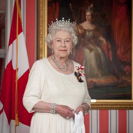 Queen Elizabeth II is dressed in a cream-coloured dress and wears a crown. She is holding a pair of white gloves. She is standing in front of a portrait of Queen Victoria. There is a Canada flag in the background.