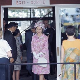 La reine Elizabeth II se tient devant un hall d’entrée ouvert. Le panneau au-dessus de l’entrée indique « Exit – Sortie ». Elle porte un manteau rose à motifs floraux, un chapeau assorti et des gants blancs et tient un sac à main blanc.