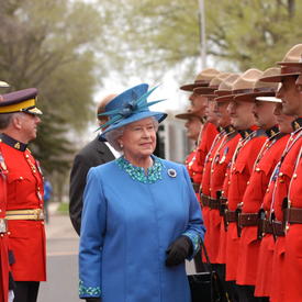 Queen Elizabeth II is dressed in a powder-blue ensemble with a matching hat. She is walking in front of a row of Royal Canadian Mounted Police officers in traditional uniforms. Two other officers look on from her right. 