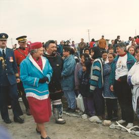 La reine Elizabeth II se déplace devant un groupe d’Inuits. Elle porte une robe de couleur fuchsia, un chapeau assorti, un pardessus bleu et des gants noirs.