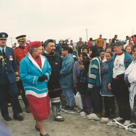 La reine Elizabeth II se déplace devant un groupe d’Inuits. Elle porte une robe de couleur fuchsia, un chapeau assorti, un pardessus bleu et des gants noirs.