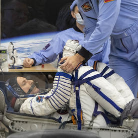 David Saint-Jacques is in his space suit and is crouched into a seat.  He is being examined by a man in a light blue jumpsuit and face mask prior takeoff.  