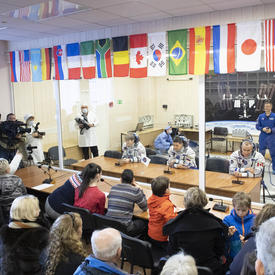 Anne McClain, Oleg Kononenko and David Saint-Jacques sit in front of microphones. They are separated from the crowd by large glass windows.  The crowd is composed of family members and media.