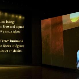 Sur un mur, une vidéo joue, sur un autre mur, les mots « Tous les êtres humains naissent libres et égaux en dignité et en droits" sont écrits ». 