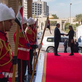 Governor General Julie Payette shakes a man's hand in the distance at the end of a red carpet while three guards dressed in red and wearing a white hat line up to the left of the carpet.