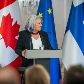 Governor General Simon speaking into a microphone at a podium. Behind her, in order from left to right, are the flags of Canada, Finland and the European Union.  