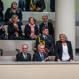 Governor General Simon standing on a balcony in Finland’s Parliament. Several parliamentarians are seated around her. They are clapping.