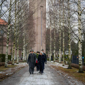 La gouverneure générale Mary Simon marche sur un chemin de gravier dans un cimetière. Un homme en uniforme marche à côté d’elle et un groupe de personnes marche derrière eux. Derrière le groupe se trouve une grande tour d’horloge en pierre.