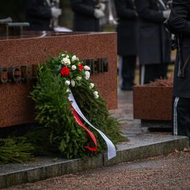 Une couronne de fleurs rouges et blanches est déposée devant un monument. Un groupe de personnes en uniforme militaire se tient autour du monument.