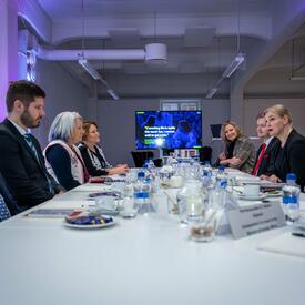 La gouverneure générale Mary Simon est assise à une grande table avec d’autres personnes. Une télévision est posée au bout de la table et une présentation y est diffusée.