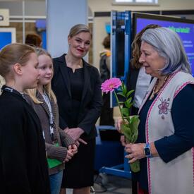 La gouverneure générale Mary Simon tient une fleur rose et s’adresse à deux écoliers. Deux femmes se tiennent derrière eux.