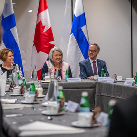 Governor General Simon sitting at the head of a large conference table, in between two other people. Behind her are two flags of Canada and two flags of Finland. Two other people sitting at the table have their backs to the camera.